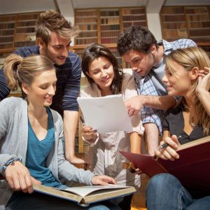 A group of five young adults sitting on the floor of a library, engaged in a discussion over papers and books.